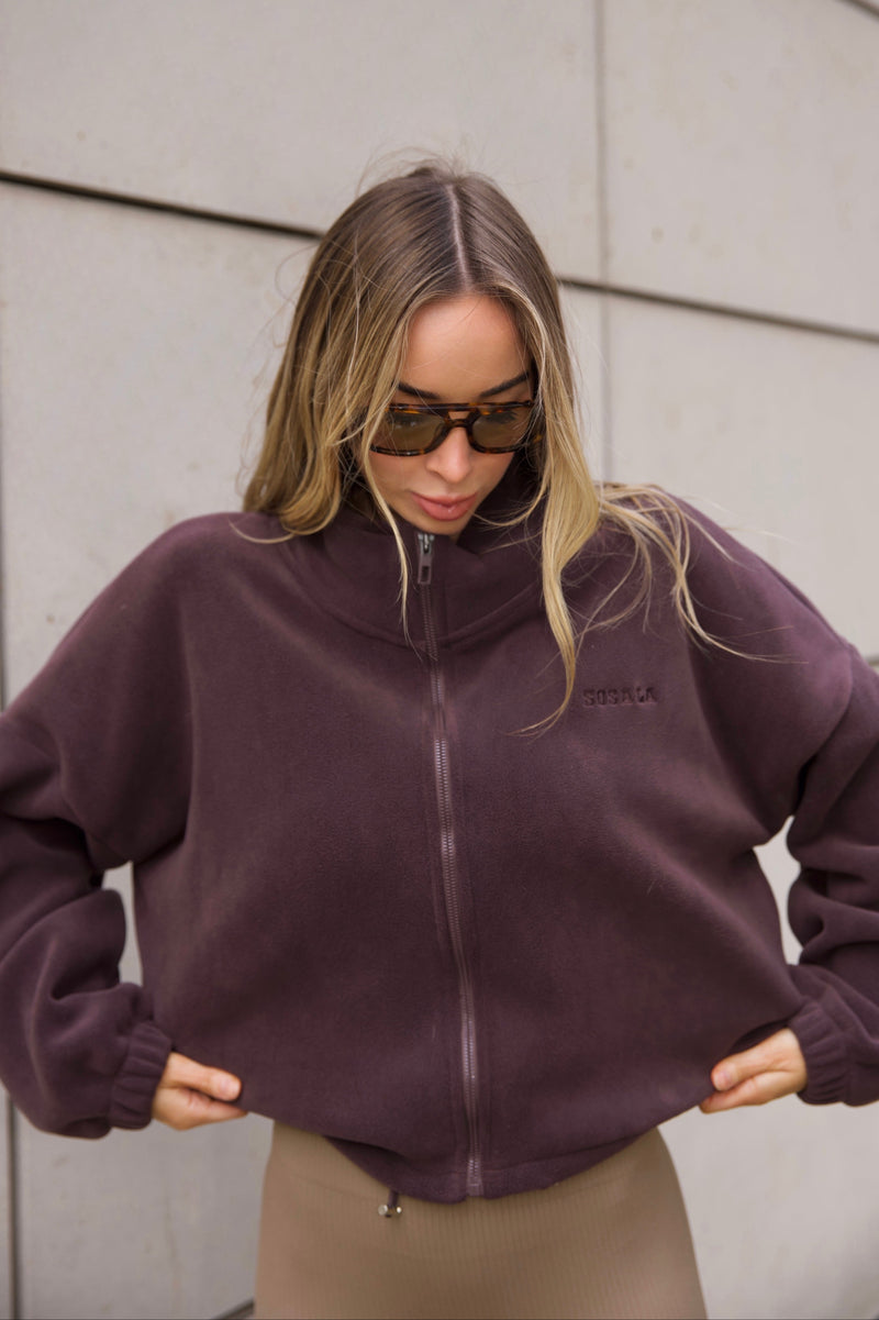 Woman wearing a dark brown fleece  jacket with a sosala brand logo and light brown leggings against a light-colored wall.