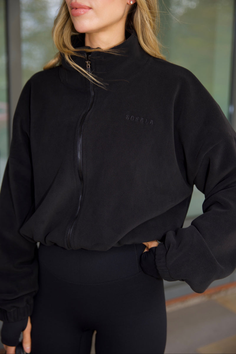 Person wearing a black fleece jacket with a sosala brand logo and black leggings, standing indoors.