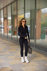 Woman in black outfit sosala matching set or zip top and leggings walking on a sidewalk with glass building reflections.