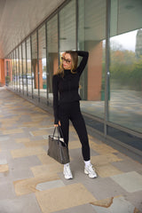Woman in black sosala matching outfit of zip top and leggings with white sneakers holding a bag, standing on a sidewalk.
