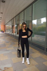 Woman in black sosala matching outfit of bralet, zip top and leggings standing on a sidewalk with glass building in the background