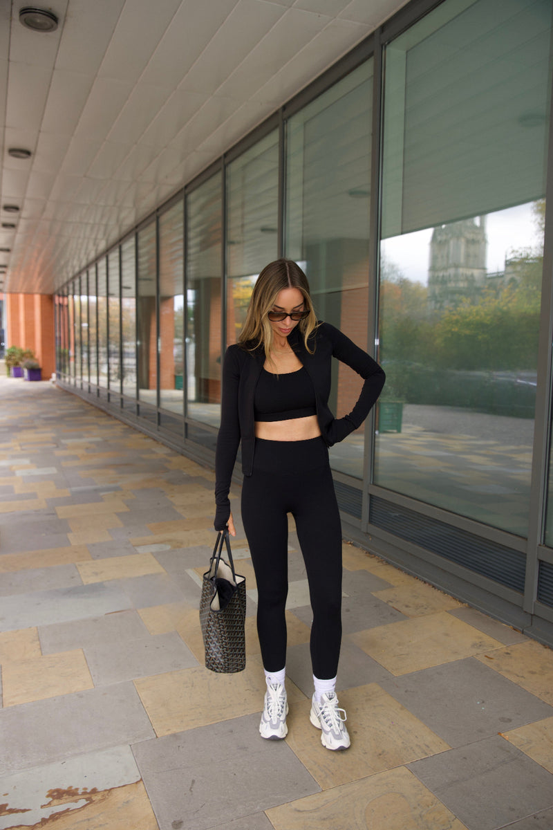 Woman in black sosala matching outfit of bralet, zip top and leggings standing on a sidewalk with glass building in the background
